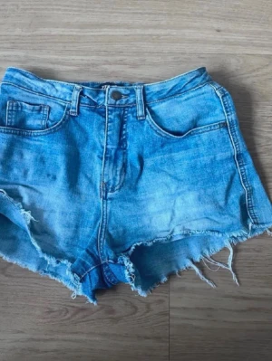 Blå jeansshorts med fransig kant - Säljer ett par ljusblå jeansshorts med fransig, rå kant och klassisk femficksmodell. Shortsen har bälteshällor och knappgylf. Perfekta för sommaren och ger en avslappnad vibe. Ok skick.