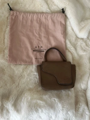 Brun miniväska från ATP Atelier - Snygg brun miniväska från ATP Atelier i slätt läder. Väskan har ett unikt lock med asymmetrisk kant och ett kort handtag upptill. Kommer med original dustbag i rosa tyg. Perfekt för dig som gillar stilrena accessoarer.