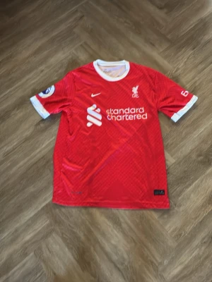 Liverpool matchtröja röd Nike - Säljer en röd Liverpool matchtröja från Nike med vita detaljer och klubbmärke på bröstet. Tröjan har Premier League-märke på ärmen, sponsorlogga framtill och är tillverkad i ett lätt, ventilerande material. Perfekt för fotbollsträning eller att bära på matchdag.xxl men sitter som l/xl oanvänd 