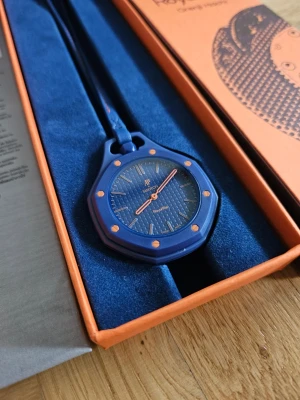 Blå Swatch Royal Pop Orenji Hachi Klocka - Säljer en blå Swatch Royal Pop klocka i samarbete med Audemars Piguet. Klockan har en rund urtavla med orange detaljer och blått band. Levereras i originalförpackning med tillhörande manualer och påse. Perfekt för dig som gillar unika accessoarer.