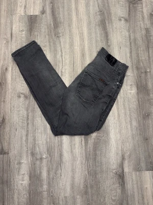 Nudie - Snygga mörkgrå nudie jeans med rak passform och klassisk femficksdesign. Jeansen har subtila slitningar och är tillverkade i ett mjukt tyg. 