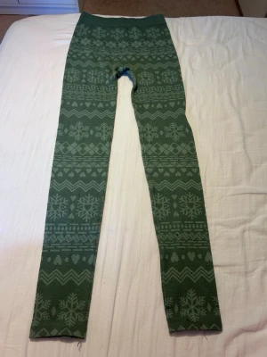 Gröna leggings med vintermönster - Säljer ett par gröna leggings med snyggt vinterinspirerat mönster av snöflingor, hjärtan och zigzag. De är stretchiga och har hög midja, perfekta för att hålla dig varm och bekväm. Materialet känns mjukt och passar bra till en chill look.