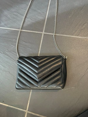 Svart quiltad axelväska med kedja - Snygg svart axelväska i quiltat mönster med silvrig kedjerem. Väskan har ett lock med magnetknäppning och ett innerfack med dragkedja. Perfekt storlek för det viktigaste och ger en trendig look till din outfit.