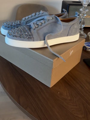 Louboutin skor  - Snygga ljusblå sneakers i mocka från Christian Louboutin med silverfärgade nitar på tån och klassisk röd sula. Skorna har vita sulor och matchande blå skosnören. Perfekt för dig som vill sticka ut med en exklusiv och edgy stil. Använt 1 gång 
