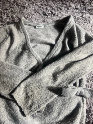 Grå stickad omlottkofta Kappahl - Mysig grå stickad omlottkofta från Kappahl med knytband i midjan och djup V-ringning. Koftan har lång ärm och är tillverkad i ett mjukt, fluffigt material som känns skönt mot huden. Perfekt för lager på lager och chill dagar.