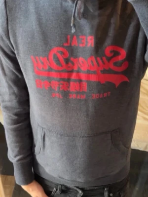 Mörkgrå Superdry hoodie med tryck - Snygg mörkgrå hoodie från Superdry med rött tryck på bröstet. Klassisk känguruficka framtill och huva . Perfekt för dig som gillar streetwear och vill ha en bekväm och stilren look.