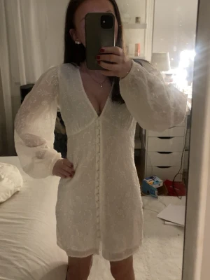 Vit kort klänning med puffärmar - Söt vit kort klänning med v-ringning, puffiga långärmar och knappar framtill. Klänningen har ett diskret blommigt mönster och är tillverkad i ett tunt, lätt material som ger en romantisk känsla. Perfekt för sommarens alla tillfällen. ” En liten fläck på axeln på sista bilden ” 