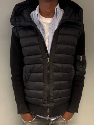 Moncler cardigan - Snygg svart moncler cardigan med stickade ärmar och stor vadderad huva. Jackan har dragkedja framtill, ficka med dragkedja på ena ärmen och quiltad front. Perfekt för kalla dagar och ger en modern look.