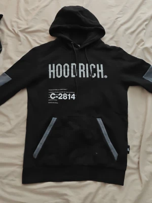 Hoodrich hoodie - Säljer en svart hoodie från HOODRICH med stora vita tryck fram och bak. Framtill står det HOODRICH och C-2814, baktill finns en världskarta och HOODRICH-loggan. Hoodien har grå detaljer på fickor, ärmar och nederkant samt en stor huva och känguruficka.