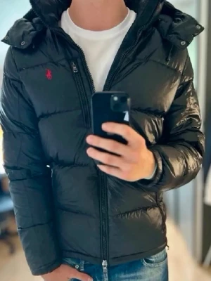 Svart dunjacka från Polo Ralph Lauren - Säljer en svart dunjacka från Polo Ralph Lauren med röd logga på bröstet. Jackan har huva, dragkedja framtill och är tillverkad i glansigt material. Perfekt för kalla vinterdagar och riktigt snygg till jeans eller andra outfits.