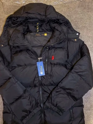 Svart dunjacka från Polo Ralph Lauren - Svart dunjacka från Polo Ralph Lauren med huva och dragkedja framtill. Jackan har två sidofickor med dragkedja och den klassiska röda logotypen broderad på bröstet. Materialet är glansigt och puffigt, perfekt för kalla dagar.