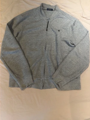 Grå ziptröja från Polo Ralph Lauren - Snygg grå ziptröja från Polo Ralph Lauren med diskret logga på bröstet. Tröjan har lång ärm, ribbade muddar och dragkedja framtill. Perfekt för lager på lager och enkel att matcha med olika outfits. Inga märkbara tecken på slitage.