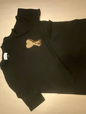 Svart t-shirt Ami Paris med brunt hjärtmärke - Snygg svart t-shirt med ett brunt hjärtmärke och bokstaven A broderat på bröstet. Klassisk rund hals och korta ärmar. Perfekt till jeans eller shorts för en clean och stilren look.