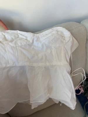 Zara tröja - Säljer en vit korsettopp med tunna axelband och snörning i sidorna. Toppen har markerade sömmar och en lätt volang nedtill för en feminin look. Perfekt att styla med jeans eller kjol för en trendig outfit.