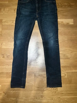 Mörkblå jeans från Nudie Jeans - Säljer ett par mörkblå jeans från Nudie Jeans med snygga slitningar och kontrastsömmar. Jeansen har en smal passform och klassisk femficksdesign. Materialet är denim och färgen är djup indigo med lätt tvättade detaljer. Lean Dean(W31)(L32)