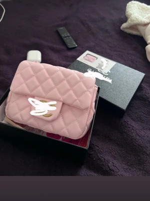 Ljusrosa quiltad handväska - Söt ljusrosa handväska med quiltat mönster och guldfärgat vridlås. Väskan har en kompakt och rektangulär form, perfekt för det viktigaste. Tillverkad i ett mjukt material som ger en lyxig känsla.