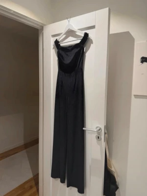 Svart off shoulder jumpsuit - Snygg svart jumpsuit med off shoulder-design och vida ben. Sitter tajt upptill och har en smickrande passform. Perfekt för dig som vill ha en stilren och trendig look. Materialet ser ut att vara mjukt och följsamt, troligen polyester eller liknande. Glittrig 