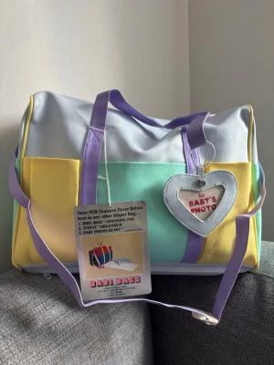 Pastellfärgad skötväska med hjärtdetalj - Säljer en stor och rymlig skötväska i pastellfärger med lila handtag och axelrem. Väskan har fack på sidorna, en hjärtformad detalj för foto och etikett framtill. Materialet är konstläder i gult, mintgrönt och ljusblått. Perfekt för att organisera babyprylar. Om du öppnar facket i botten så finns en skötbädd som du enkelt viker ut. 