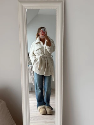 Vit teddyjacka med knytskärp - Mysig vit teddyjacka med knappar framtill och knytskärp i midjan. Jackan har en avslappnad passform och är tillverkad i fluffigt teddy-material som håller dig varm och trendig.