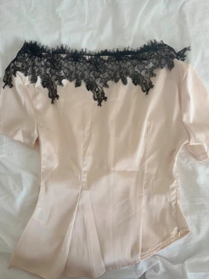 Ljusbeige  blus med svart spets - Snygg ljusbeigh blus med korta ärmar och dekorativ svart spets längs offshoulder-kragen. Tillverkad i ett glansigt, mjukt material som ger en elegant look. Perfekt för dig som vill ha något extra till outfiten.