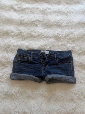 Blå jeansshorts från Abercrombie&fitch  - Snygga blå jeansshorts med uppvikta ben från Abercrombie&fitch. Shortsen har en liten broderad logga på fickan och kontrastsömmar. Perfekta för varma dagar och ger en avslappnad look.