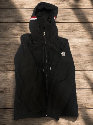 Svart moncler vindjacka med huva - Svart vindjacka med dragkedja och huva. Jackan har en vit och röd detalj på huvan samt en rund logotyp på bröstet. Tillverkad i lätt material, perfekt för blåsiga dagar. Snygg och enkel design med elastiska muddar vid ärmsluten.