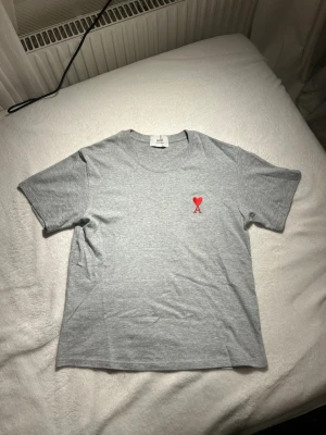 Grå t-shirt med rött hjärta från Ami - Säljer en stilren grå t-shirt från Ami med ett litet rött hjärta och bokstaven A broderat på bröstet. Klassisk rund halsringning och korta ärmar. Perfekt till jeans eller shorts för en clean look.