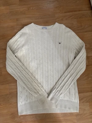Stickad tröja gant vit - Säljer en klassisk vit kabelstickad tröja från GANT i storlek M. Tröjan har rund halsringning, ribbade muddar och diskret GANT-logga broderad på bröstet. Perfekt för dig som gillar stilrena och tidlösa plagg i mjukt material.