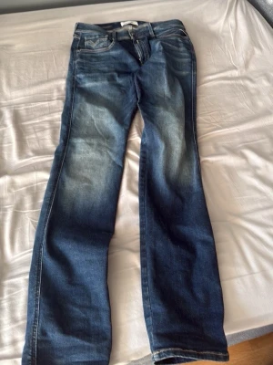 Blå jeans från Replay Anbass - Säljer ett par snygga blå jeans från Replay, modell Anbass. Jeansen har en klassisk femficksdesign, raka ben och coola slitningar framtill. Tillverkade i mjukt denimtyg som sitter skönt och passar till det mesta. Perfekta för dig som gillar en avslappnad men stilren look.