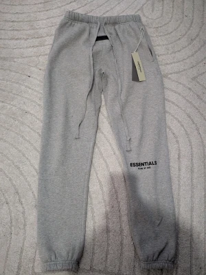 Gråa Essentials mjukisbyxor -         LETAR EFTER BUD BUDET: 0  (vuxen storlek XS)                                                                        Säljer ett par gråa mjukisbyxor från Fear of God Essentials. Byxorna har resår i midjan och benslut, dragsko i midjan och tryckt logga på benet. Tillverkade i mjukt material som är perfekt för chill dagar. Snygg och enkel design med streetwear-vibe.