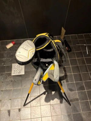 Golfset med bag och klubbor - Säljer ett golfset med svart och gul bärbag samt flera klubbor, inklusive järn 7, S-wedge och en driver. Bagen har flera fack och axelremmar för enkel transport. Perfekt för dig som vill börja spela golf eller behöver ett extra set.
