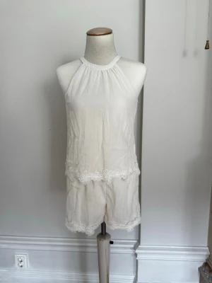 Ärmlös jumpsuit med spetsdetaljer - Obs! Barnstorlek 156 men passar XXS. Säljer en söt cremefärgad jumpsuit med halterneck och fina spetsdetaljer vid midjan och bensluten. Modellen är ärmlös och har en avslappnad passform, perfekt för sommardagar. Materialet ser ut att vara lätt och luftigt, vilket gör den skön att bära.