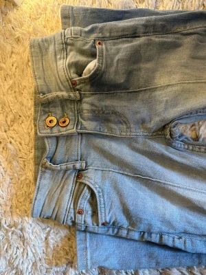 Ljusblå jeans med dubbla knappar - Säljer ett par ljusblå jeans med raka ben och dubbla knappar i midjan. Jeansen har klassiska fem fickor och kontrastsömmar. Materialet är denim i bomull och de har en avslappnad passform som passar till många olika stilar.