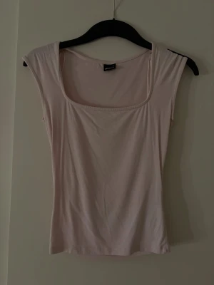 Ljusrosa  topp med fyrkantig ringning - Säljer en ljusrosa topp med fyrkantig ringning och ärmlös design. Toppen har en slim passform och är tillverkad i mjukt bomullsmaterial. Perfekt att styla med jeans eller kjol för en enkel och fräsch look.