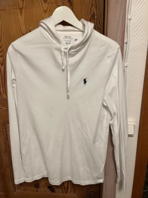 Vit hoodie från Polo Ralph Lauren - Säljer en stilren vit hoodie från Polo Ralph Lauren med klassisk huva och dragsko. Tröjan har lång ärm och den ikoniska mörkblå loggan broderad på bröstet. Tillverkad i mjukt bomullsmaterial som känns skönt mot huden. Perfekt för en clean och avslappnad look.