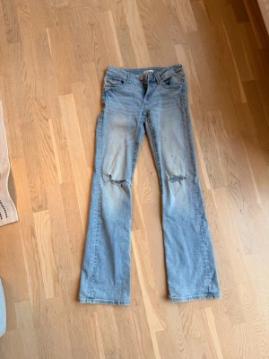 Ljusblå slitna jeans från Gina Tricot - Ljusblå jeans från Gina Tricot med raka ben och slitningar över knäna. Klassisk femficksmodell med knapp och dragkedja framtill. Perfekta för en avslappnad och trendig look.