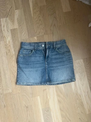 Blå jeanskjol från H&M - Säljer en klassisk blå jeanskjol från H&M i kort modell. Kjolen har fem fickor, knapp och dragkedja framtill samt bälteshällor. Perfekt till vardags för en avslappnad och trendig look.