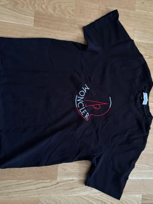 Svart Moncler t-shirt med stor logga - Snygg svart t-shirt från Moncler med stor rund logga i vitt och rött på bröstet. Klassisk passform och korta ärmar. Tillverkad i mjuk bomull som känns skön mot huden. Perfekt för dig som gillar stilrena och exklusiva plagg.