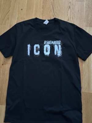 Svart ICON t-shirt med tryck - Snygg svart t-shirt med stort vitt 'ICON' tryck på bröstet och Dsquared2-inspirerad text ovanför. Klassisk rund hals och korta ärmar. Tillverkad i mjuk bomull som känns skön mot huden. Perfekt för dig som gillar streetwear och tydliga statements.