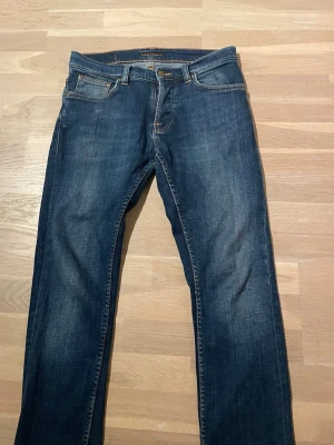 Mörkblå jeans från Nudie Jeans - Säljer ett par mörkblå jeans från Nudie Jeans med klassisk femficksmodell och orangea sömmar. Jeansen har raka ben och är tillverkade i slitstark denim. Snygga detaljer på bakfickorna och patch med logga bak i midjan.