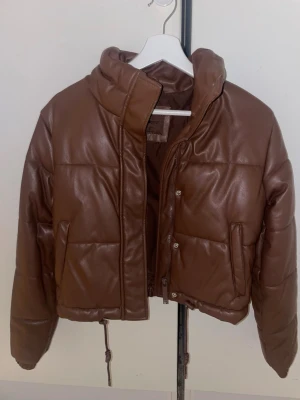 Brown leather jacket  - Säljer en brun pufferjacka från FBSISTER i storlek S. Jackan har hög krage, dragkedja och tryckknappar framtill samt dragsko i nederkant. Materialet är glansigt och ger en cool look. Perfekt för kalla dagar när du vill vara både varm och snygg. Priset kan diskuteras 