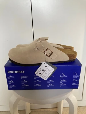 Beige Birkenstock tofflor i mocka - Säljer ett par snygga beige tofflor från Birkenstock i mjuk mocka med justerbart spänne i metall. Klassisk slip-in modell med korksula och ergonomisk fotbädd för extra komfort. Perfekta för chill dagar hemma eller när du vill ha det bekvämt.