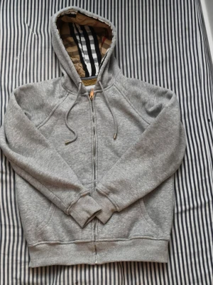 Grå zip hoodie från Burberry - Säljer en grå zip hoodie från Burberry med klassiskt rutigt foder i huvan och dragsko. Hoodien har fickor framtill, ribbade muddar och är gjord i mjukt material. Perfekt för dig som vill ha en stilren och lyxig look.