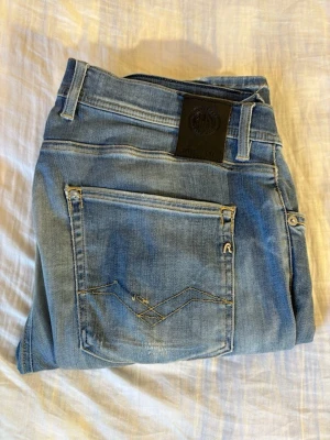 Ljusblå raka jeans med broderad ficka Anbass - Säljer ett par ljusblå jeans med raka ben och snygga broderidetaljer på bakfickan. Jeansen har klassisk femficksdesign, bälteshällor och en svart patch bak i midjan. Perfekta till en avslappnad stil och passar till det mesta. Dess är ett par replay anbass 