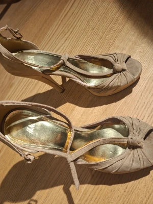 Beige pumps med knut och T-rem - Snygga beige pumps med hög klack och T-rem. Skorna har en dekorativ knut framtill och är gjorda i mockaimitation. Guldfärgad innersula ger en extra touch. Perfekta för dig som gillar stilrena och trendiga skor med öppen tå och justerbart spänne vid ankeln.