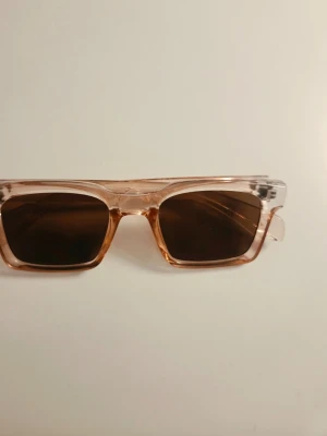 Rektangulära solglasögon i transparent rosa - Snygga solglasögon med rektangulära bågar i transparent rosa plast och mörka glas. De har en modern och trendig look som passar perfekt till soliga dagar. Lätta att matcha med olika outfits och ger en cool vibe.