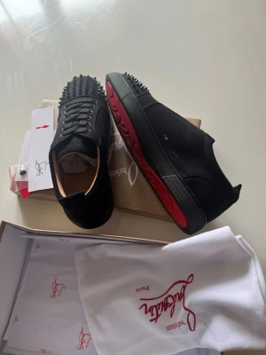 Svarta sneakers med nitar och röd sula - Snygga svarta sneakers från Christian Louboutin med mattsvarta nitar på tån och klassisk röd sula. Skorna har en låg profil, svarta skosnören och är tillverkade i mocka. Perfekta för dig som vill sticka ut med en edgy och exklusiv stil.