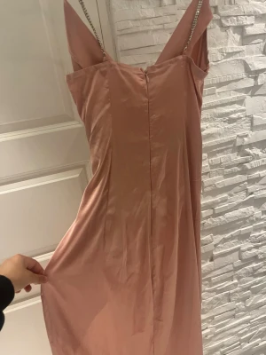 Lång rosa klänning med kedjeband - Elegant långklänning i ljusrosa satin med smala kedjeband som axelband. Klänningen har en draperad rygg och en slät framsida, vilket ger en lyxig och trendig look. Perfekt för dig som vill sticka ut med en stilren och modern vibe.