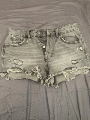 Gråa  jeansshorts - Säljer ett par gråa jeansshorts med slitna detaljer och fransig kant. Shortsen har klassisk femficksdesign, bälteshällor och knappar framtill. Perfekta för en avslappnad och trendig sommarlook.