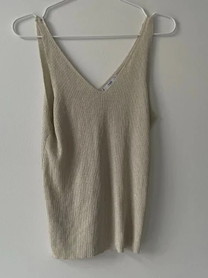 Guld ribbad linnetopp med v-ringning - Säljer en stilren beige linnetopp med v-ringning och ribbad struktur. Toppen är ärmlös och har en enkel, clean look som passar perfekt till sommaren. Materialet känns mjukt och följsamt mot huden.